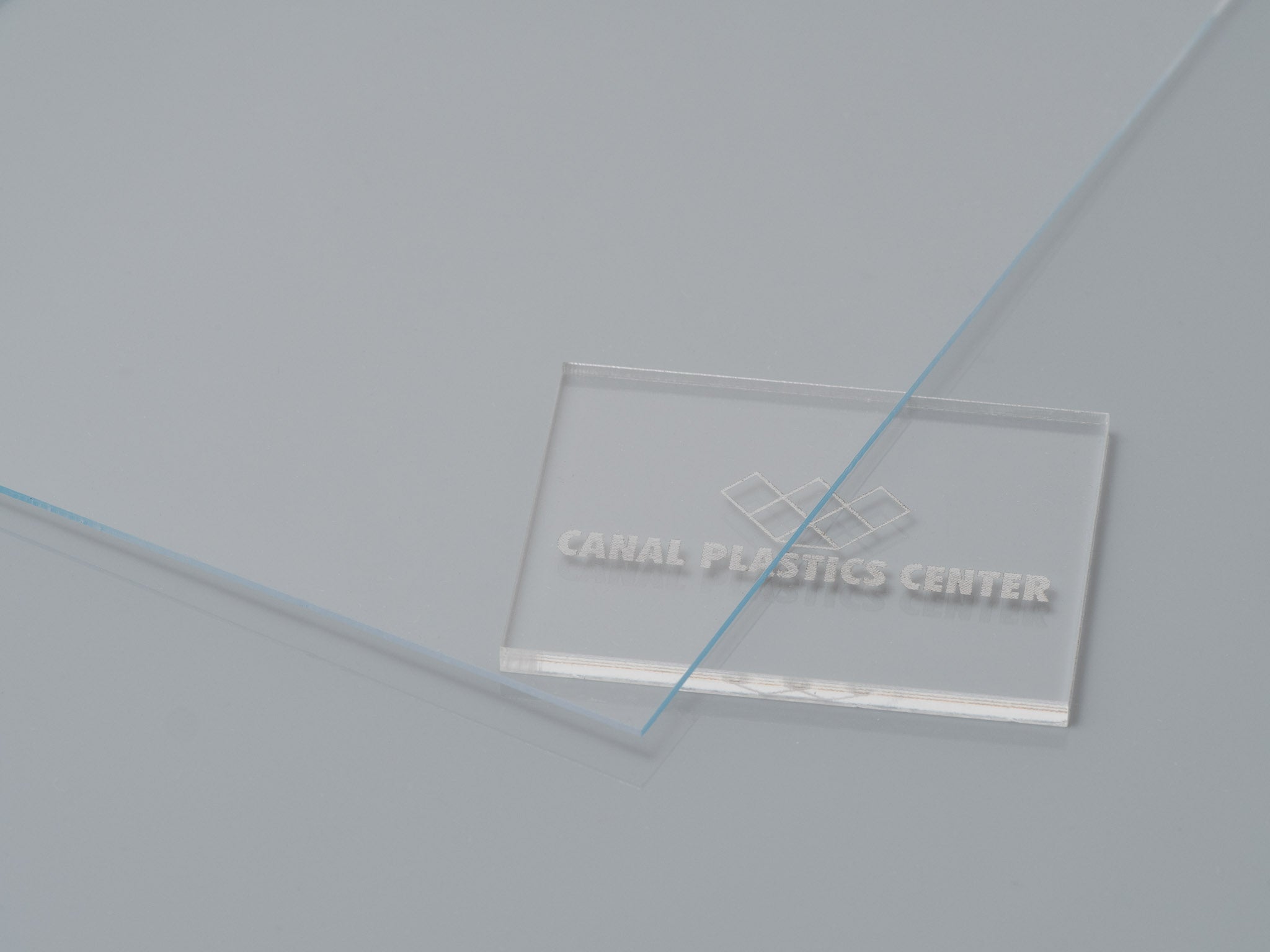 A clear acrylic film sample with 'CANAL PLASTICS CENTER' branding lying on a grey surface. 

Canal Plastics Center Clear acrylic film as a versatile material with numerous applications, offering a combination of weather resistance, moldability, and transparency in automotive, building, and display applications.